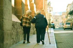 Duas pessoas idosas caminhando juntas pela calçada, uma delas com auxílio de bengala.