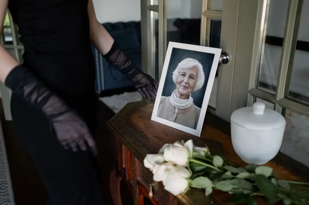 Pessoa vestida de preto ao lado de retrato de uma idosa, flores brancas e urna funerária.