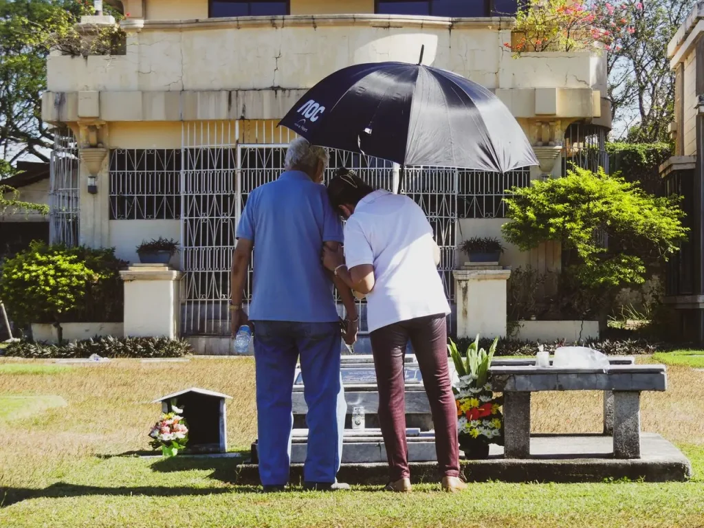 Duas pessoas de costas sob um guarda-chuva diante de um túmulo com flores em um cemitério.