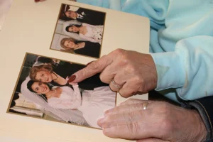Pessoa idosa folheando um álbum de fotografias e apontando para uma foto de casamento, onde aparecem uma noiva, uma mulher mais velha e um homem de terno.