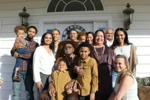 Família multigeracional reunida em frente à entrada de uma casa, com adultos, idosos e crianças sorrindo para uma foto em grupo ao ar livre.