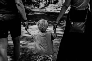 Criança caminhando de mãos dadas com dois adultos em uma trilha arborizada.