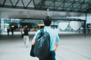 Homem de costas, vestindo camisa azul-clara e carregando mochila preta, caminhando por um espaço amplo e moderno, com estrutura metálica e outras pessoas ao fundo.