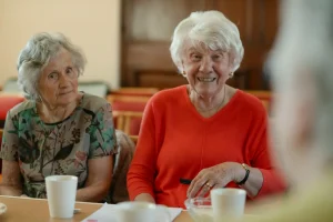 Duas mulheres idosas sentadas à mesa, uma com blusa floral e outra com blusa vermelha, participam de uma conversa em ambiente interno.
