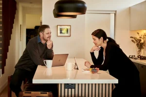 Homem e mulher conversam enquanto consultam um notebook e um tablet na bancada de uma cozinha residencial.