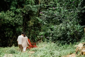 Casal caminhando por um jardim cercado de árvores e vegetação densa, próximo a cadeiras de madeira vermelhas em meio à natureza.